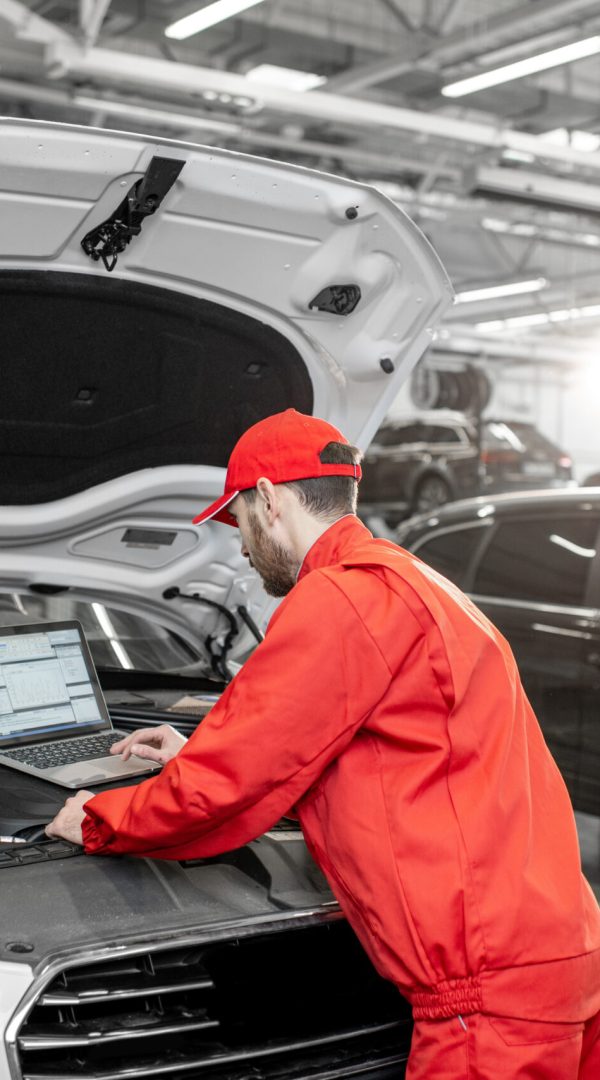 Handsome auto mechanic in red uniform doing engine diagnostics with computer in the car service
